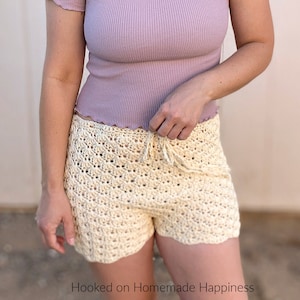 May include: A woman wearing a light purple ribbed crop top with a scalloped hem and cream crochet shorts. The shorts have a drawstring waist and a scalloped hem.
