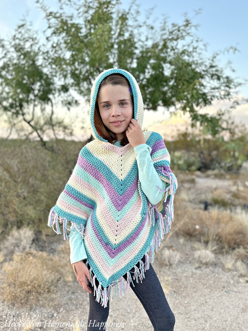 May include: A colorful, hooded crochet poncho in pastel shades of blue, purple, and cream. The poncho features a chevron pattern and fringe along the bottom edge. The hood is trimmed in a contrasting blue color.