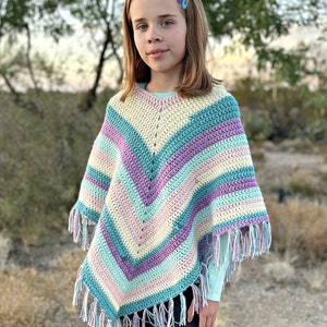 May include: A colorful, handmade crochet poncho with a V-neck design. The poncho features stripes of pastel colors including blue, purple, pink, and cream, with fringe along the bottom edge. The words "Hooked on Homemade Happiness" are visible in the bottom left corner.