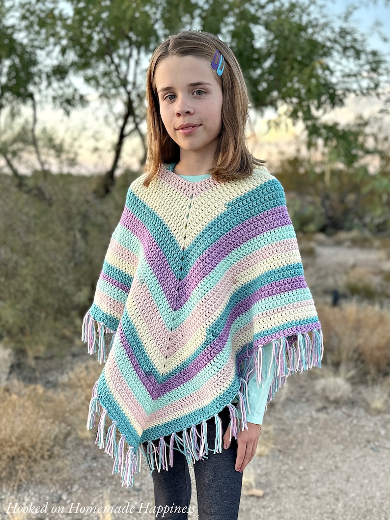 May include: A colorful, handmade crochet poncho with a V-neck design. The poncho features stripes of pastel colors including blue, purple, pink, and cream, with fringe along the bottom edge. The words "Hooked on Homemade Happiness" are visible in the bottom left corner.