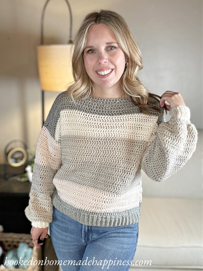 Puede incluir: Un su&eacute;ter de crochet en crema, gris y verde claro. El su&eacute;ter tiene cuello redondo y mangas largas abullonadas. El texto "hookedonhomemadehappiness.com" es visible en la parte inferior de la imagen.