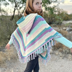 May include: A colorful, hooded crochet poncho with fringe. The poncho features stripes of pastel colors including blue, purple, pink, and yellow. The fringe is a mix of white and pastel colors. The poncho is worn by a person.