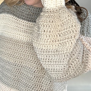Puede incluir: Un su&eacute;ter de ganchillo hecho a mano con un patr&oacute;n de rayas grises, crema y rosa claro. El su&eacute;ter presenta un pu&ntilde;o acanalado color crema y una puntada suelta y texturizada en el cuerpo y las mangas. Las mangas son estilo globo.