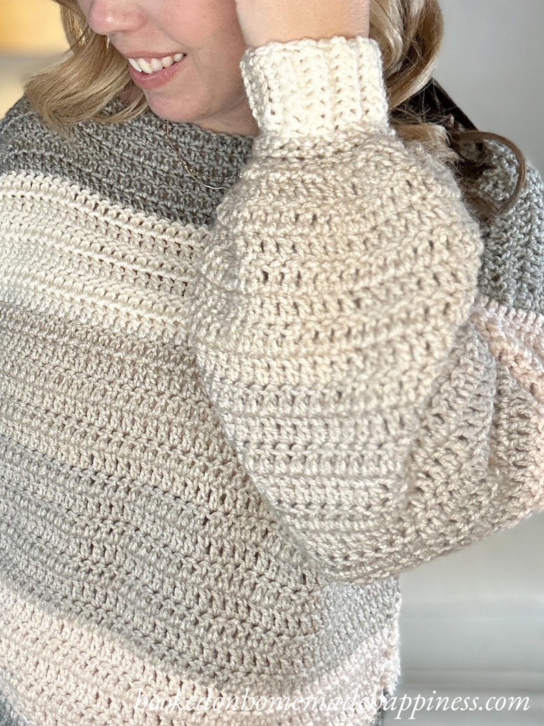 Puede incluir: Un su&eacute;ter de ganchillo hecho a mano con un patr&oacute;n de rayas grises, crema y rosa claro. El su&eacute;ter presenta un pu&ntilde;o acanalado color crema y una puntada suelta y texturizada en el cuerpo y las mangas. Las mangas son estilo globo.