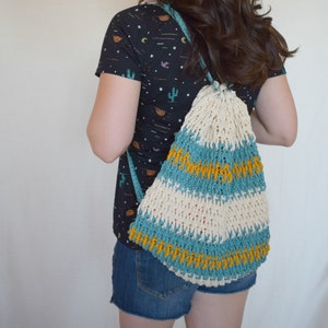 May include: A person wearing a black and white patterned shirt with a crocheted bag with blue, white, and yellow stripes. The bag is worn as a backpack.