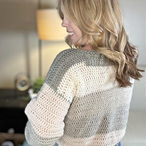 Puede incluir: Un su&eacute;ter tejido a mano en tonos grises, crema y rosa p&aacute;lido. El su&eacute;ter presenta un dise&ntilde;o a rayas con mangas largas y cuello redondo. El sitio web "hookedonhomemadehappiness.com" es visible en la esquina superior izquierda.