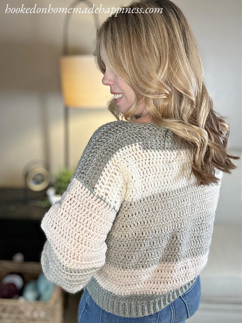 Puede incluir: Un su&eacute;ter tejido a mano en tonos grises, crema y rosa p&aacute;lido. El su&eacute;ter presenta un dise&ntilde;o a rayas con mangas largas y cuello redondo. El sitio web "hookedonhomemadehappiness.com" es visible en la esquina superior izquierda.