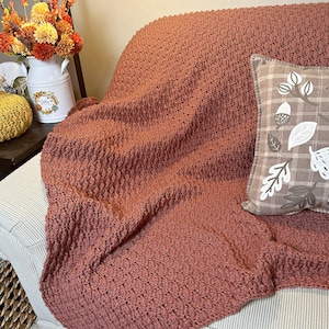 Puede incluir: Una manta de ganchillo texturizada de color óxido sobre un sofá a rayas. Un cojín decorativo con diseños de hojas y bellotas descansa sobre el sofá. Una jarra blanca con flores de otoño está cerca. El texto "hookedonhomemadehappiness" es visible.