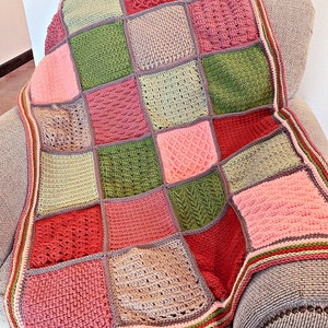 May include: A handmade crochet blanket with a patchwork design. The blanket features squares in various textures and shades of pink, green, and beige, bordered with a striped edge. It is draped over a gray armchair.