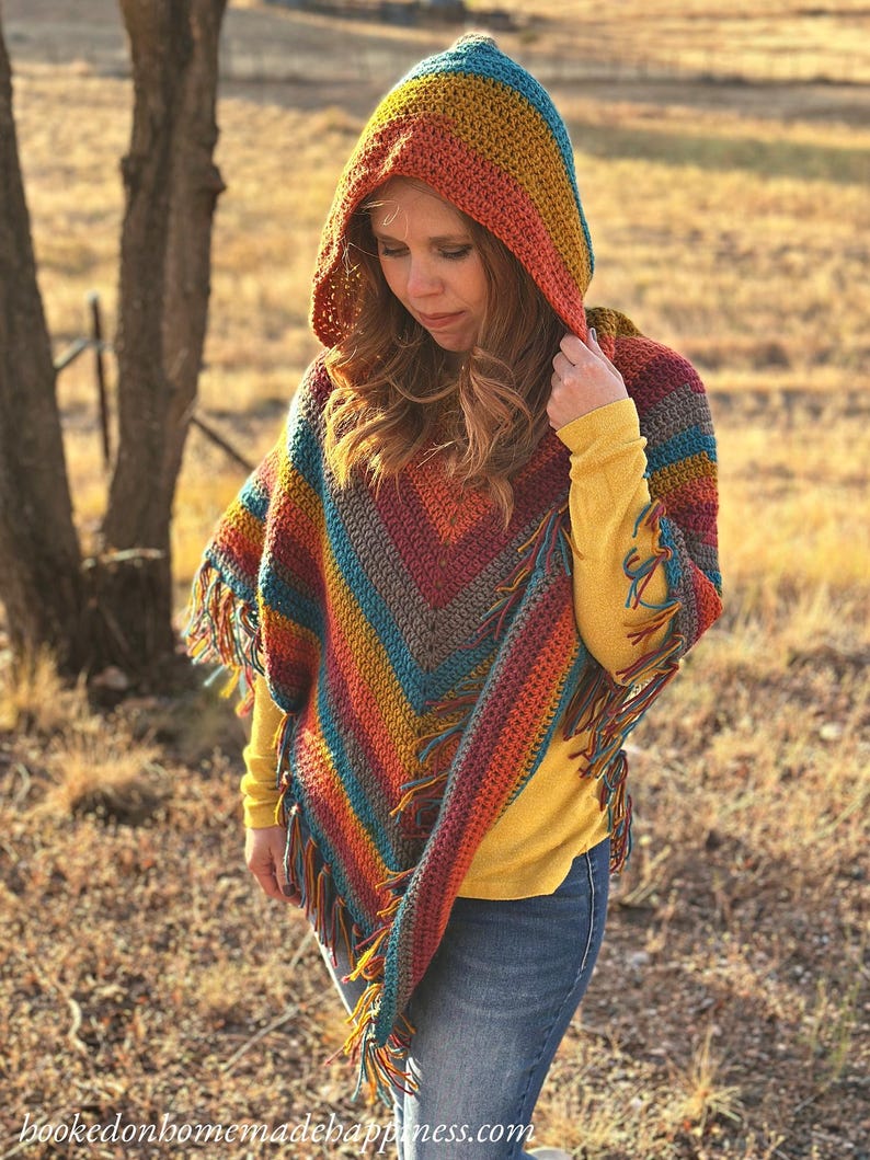 May include: A colorful, hooded poncho with a fringed hem. The crochet poncho features a vibrant array of colors, including blue, orange, yellow, and red. The text "hookedonhomemadehappiness.com" is visible at the bottom of the image.