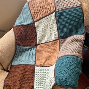 May include: A crocheted blanket with a patchwork design featuring squares in various colours and textures, including brown, beige, teal, and green. The blanket is folded and draped over a chair.