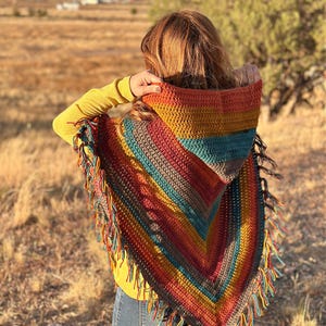 May include: A colorful, hooded crochet poncho with fringe. The poncho features a striped pattern in shades of orange, yellow, teal, and brown. The text "hookedonhomemadehappiness.com" is visible at the bottom. The person is wearing a yellow long-sleeve shirt and blue jeans.