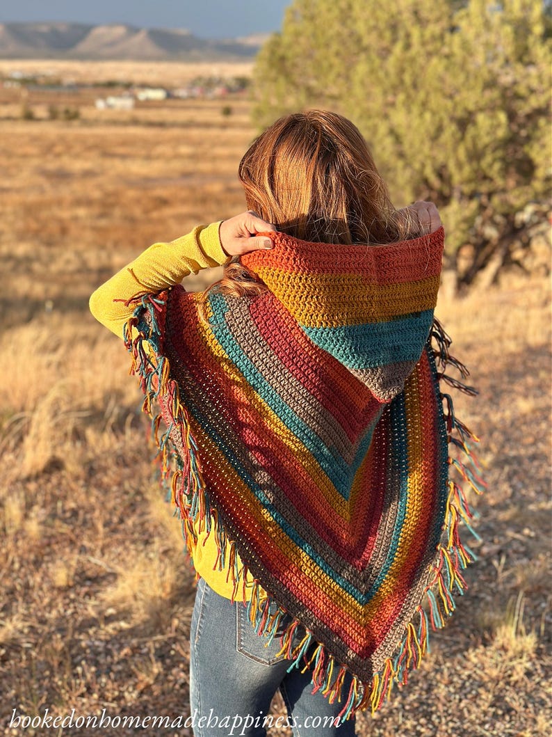 May include: A colorful, hooded crochet poncho with fringe. The poncho features a striped pattern in shades of orange, yellow, teal, and brown. The text "hookedonhomemadehappiness.com" is visible at the bottom. The person is wearing a yellow long-sleeve shirt and blue jeans.