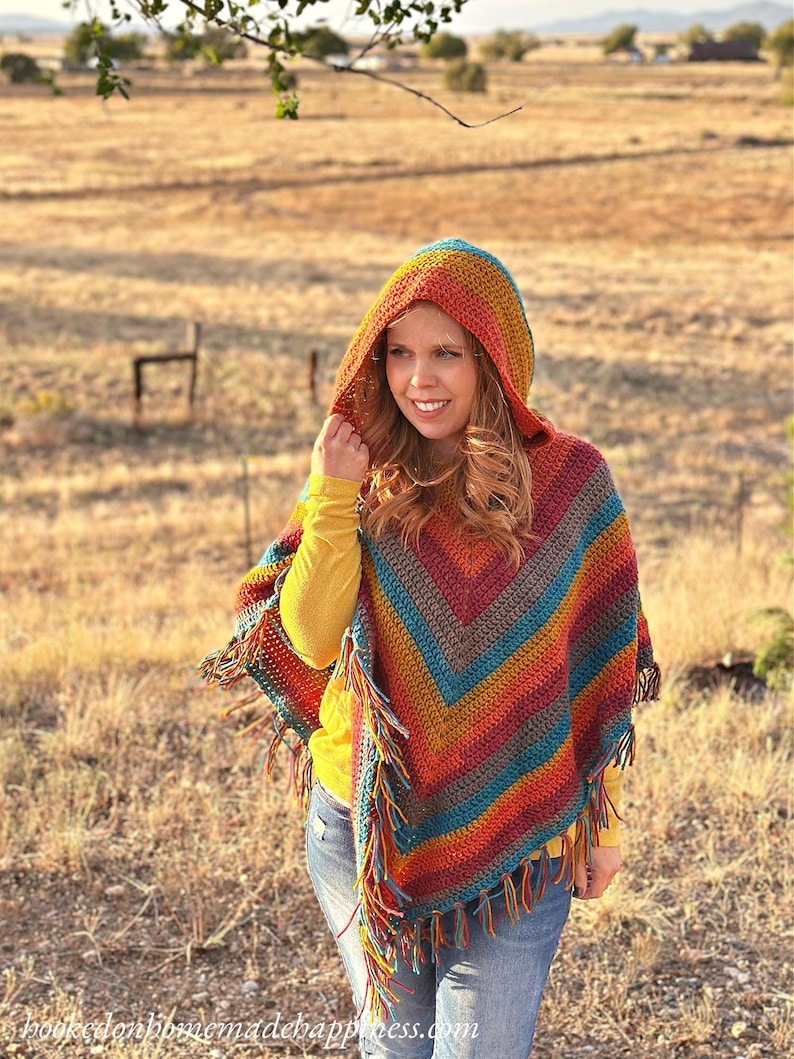May include: A colorful, hooded crochet poncho with fringe. The poncho features a striped pattern in shades of orange, red, blue, and brown. The model is wearing a yellow long-sleeve shirt and blue jeans. The website address is visible at the bottom of the image.