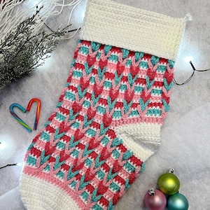 Puede incluir: Un calcetín de Navidad tejido a crochet con un puño blanco y un patrón de chevron rosa, turquesa y rojo. El calcetín está sobre una superficie blanca y esponjosa con luces navideñas y adornos.
