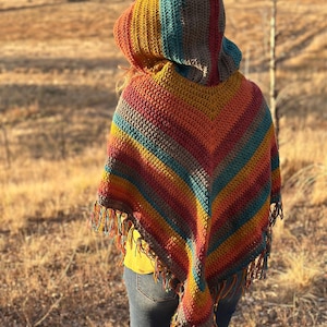 May include: A handmade crochet poncho with a hood, featuring a vibrant array of colors including red, orange, yellow, teal, and brown stripes. The poncho has a fringed hem and is worn outdoors.