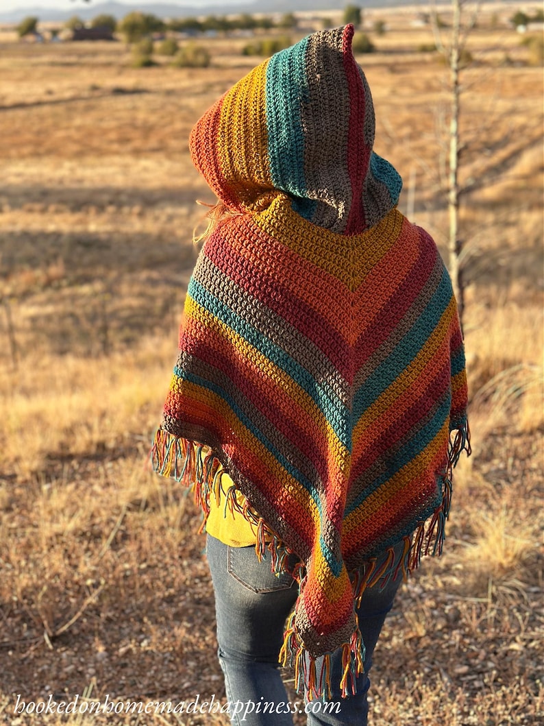 May include: A handmade crochet poncho with a hood, featuring a vibrant array of colors including red, orange, yellow, teal, and brown stripes. The poncho has a fringed hem and is worn outdoors.