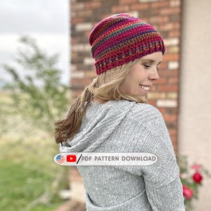 Puede incluir: Un gorro de ganchillo con rayas horizontales en tonos magenta, verde azulado, morado y naranja. El gorro tiene un borde acanalado y un patrón de puntos texturizado. El texto "PDF PATTERN DOWNLOAD" aparece en un gráfico rectangular.