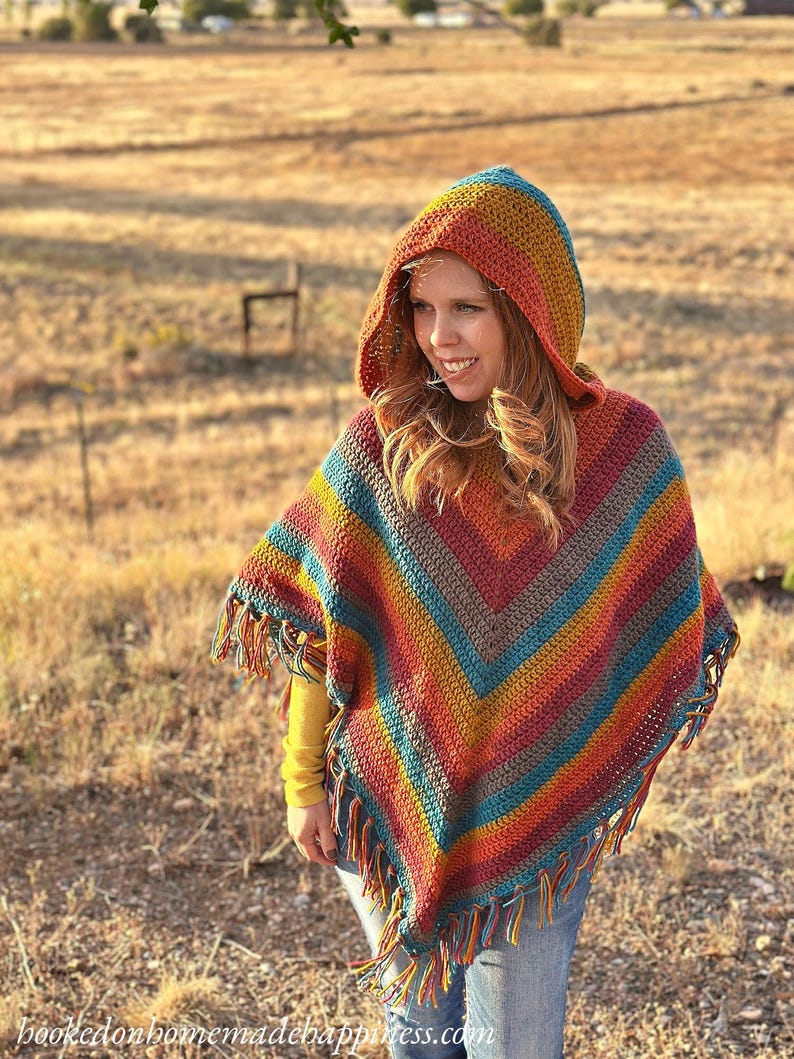 May include: A colorful, hooded crochet poncho with a fringed hem. The poncho features a striped pattern in shades of orange, yellow, teal, and burgundy. The text "hookedonhomemadehappiness.com" is visible at the bottom of the image.
