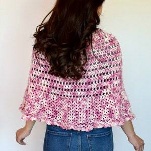 May include: A pink and white crocheted shawl with a scalloped edge. The shawl is worn by a person with long brown hair.