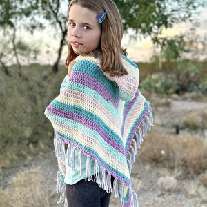 May include: A colorful, handmade crochet poncho with a hood and fringe. The poncho features stripes of blue, purple, yellow, and white. The fringe is a mix of white and light purple. The words "Hooked on Homemade Happiness" are in the bottom left corner.