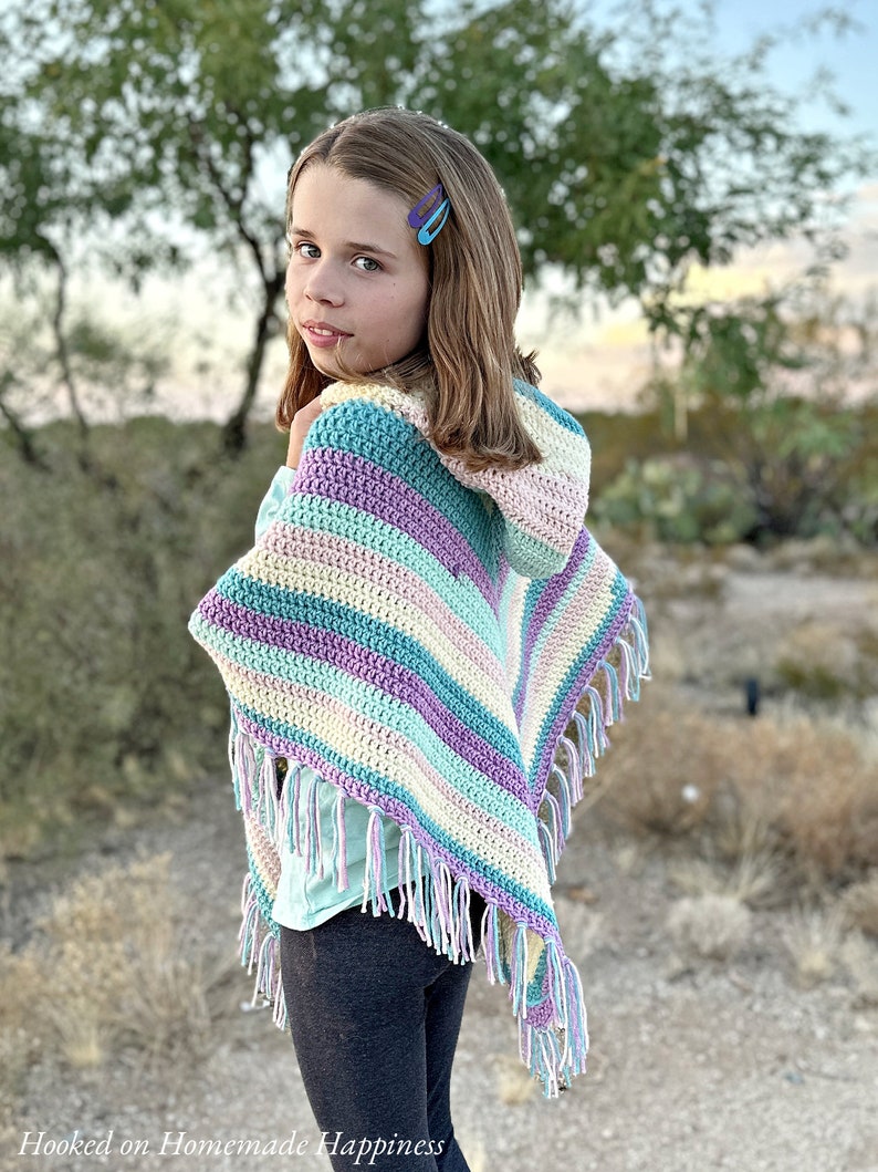 May include: A colorful, handmade crochet poncho with a hood and fringe. The poncho features stripes of blue, purple, yellow, and white. The fringe is a mix of white and light purple. The words "Hooked on Homemade Happiness" are in the bottom left corner.