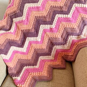 May include: A crocheted chevron blanket in shades of pink, peach, and brown. The blanket is made with a textured stitch and has a scalloped edge.