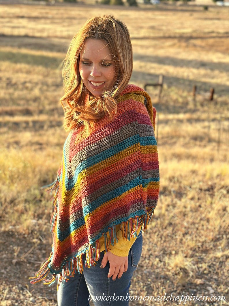 May include: A colorful, handmade crochet poncho with a hood and fringe. The poncho features stripes of red, orange, yellow, teal, and brown. The fringe is a mix of colors. The text "hookedonhomemadehappiness.com" is visible.