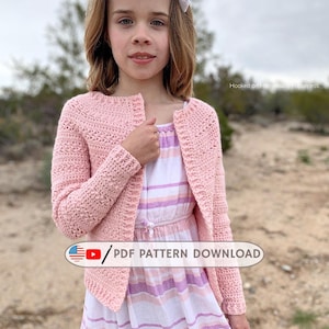 May include: A young person wearing a light pink crochet cardigan over a striped dress. The cardigan has a textured stitch pattern. The dress has a white, pink, and purple striped pattern. A white bow is in the person's hair. The image includes the text "PDF PATTERN DOWNLOAD".