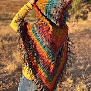 May include: A colorful, handmade crochet hooded poncho with a fringed hem. The poncho features a striped pattern in shades of orange, yellow, teal, and brown. The hood is up, and the poncho is worn over a yellow long-sleeved shirt and blue jeans.