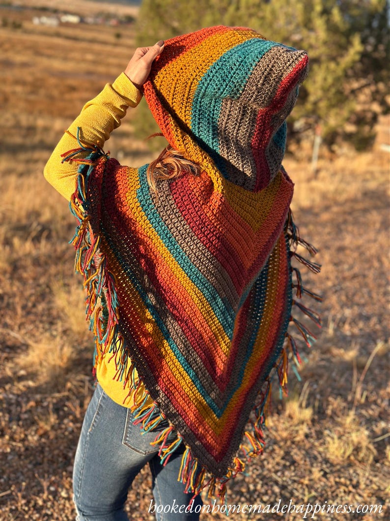 May include: A colorful, handmade crochet hooded poncho with a fringed hem. The poncho features a striped pattern in shades of orange, yellow, teal, and brown. The hood is up, and the poncho is worn over a yellow long-sleeved shirt and blue jeans.