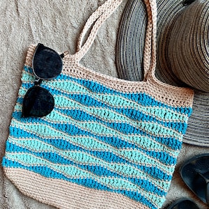 May include: A crocheted striped tote bag in beige, turquoise, and blue with two pairs of sunglasses inside. The bag is lying on a beige towel with a straw hat and black flip flops beside it.