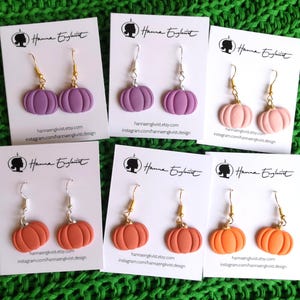Op de afbeelding: Verschillende paar pompoen oorbellen in paars, roze en oranje, gepresenteerd op witte kaarten. Elke kaart bevat de tekst "Hanna Engkvist" en social media details. De oorbellen hebben gouden of zilveren haken. De achtergrond is groen.