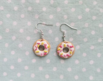 May include: A pair of donut-shaped earrings. Each earring has a tan base, white icing, and colourful sprinkles. The earrings are suspended from silver-coloured hooks. The background is a light blue fabric with white polka dots.