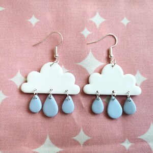 May include: A pair of dangle earrings featuring white cloud-shaped charms with three light blue teardrop-shaped rain charms hanging from each cloud by silver-toned metal jump rings and silver-toned fishhook ear wires for pierced ears.