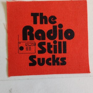 May include: Red fabric patch with black text that reads "The Radio Still Sucks" and a small black and white illustration of a radio.