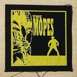 Puede incluir: Parche tejido negro y amarillo con el texto "The Mopes" en letras grandes amarillas. Una silueta de una persona con un sombrero de vaquero está de pie frente a un fondo amarillo.