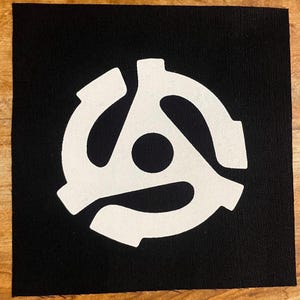 May include: A square black patch featuring a white 45 RPM record adapter design. The adapter has a circular center and three curved arms extending outwards. The design is centered on the patch.