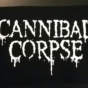 May include: Black rectangular patch with the white text "CANNIBAL CORPSE" in a dripping font. The patch is likely for a jacket or bag, representing a heavy metal band.