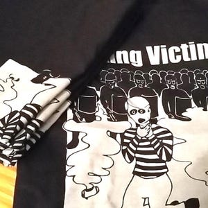 May include: Black graphic tee and matching folded cloth napkins. The tee features a black and white illustration of a person in a striped shirt, with the text "Young Victim" above a crowd of figures. The napkins have a similar design.