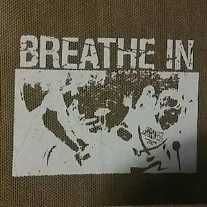 Könnte beinhalten: Ein grüner Stoff-Patch mit weißem Text, der "BREATHE IN" lautet. Der Patch zeigt eine weiße Silhouette von zwei Personen, die sich umarmen.
