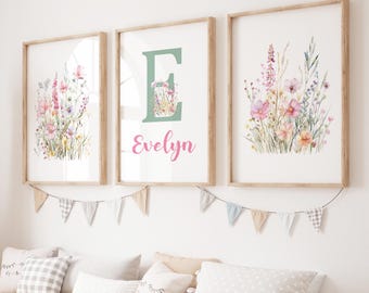 May include: Three framed floral art prints with wooden frames. The prints feature watercolor illustrations of wildflowers in pink, purple, and yellow hues. The center print has a large green letter "E" with the name "Evelyn" in pink. A garland of small flags hangs below.