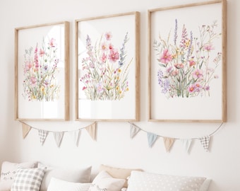 May include: Three framed floral art prints featuring watercolor paintings of wildflowers in pink, purple, and yellow hues. The prints are in light brown wooden frames and are displayed above a row of pillows and a decorative banner.