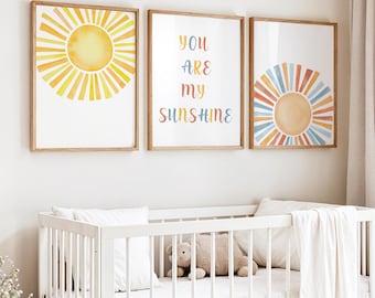 May include: Three framed watercolor art prints with a sun theme. One print features a yellow sunburst, another has the text "YOU ARE MY SUNSHINE" in colorful letters, and the third depicts a sun with orange, blue, and yellow rays. A white crib is in the foreground.