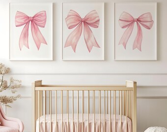 May include: Three framed watercolor prints featuring pink bows. The bows have detailed shading and flowing ribbons. The prints are displayed above a light-colored wooden crib with a pink crib skirt and a matching pink chair.