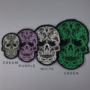 May include: Four embroidered skull patches in different colors: cream, purple, white, and green. Each skull is decorated with a lace-like design.