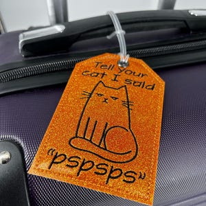 May include: Orange glitter luggage tag with a black cat illustration and the text "Tell your cat I said pspsps". The tag is attached to a purple suitcase with a white plastic loop.