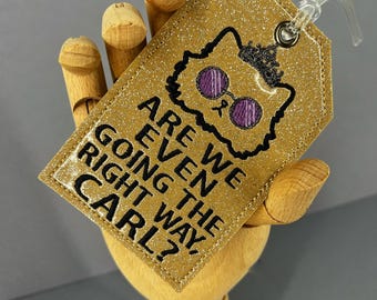 May include: A gold glitter luggage tag featuring an embroidered cat wearing a crown and purple sunglasses. The text reads, ARE WE EVEN GOING THE RIGHT WAY, CARL? The tag includes a clear plastic loop attached through a metal grommet at the top.
