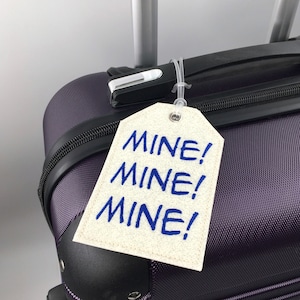 May include: A luggage tag with the word "MINE!" repeated three times in blue on a cream-colored tag. The tag is attached to a purple suitcase with a black handle.