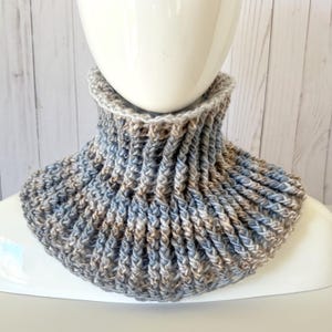 May include: A crocheted neck warmer in shades of blue and brown. The neck warmer is made with a textured stitch and has a wide, comfortable fit.